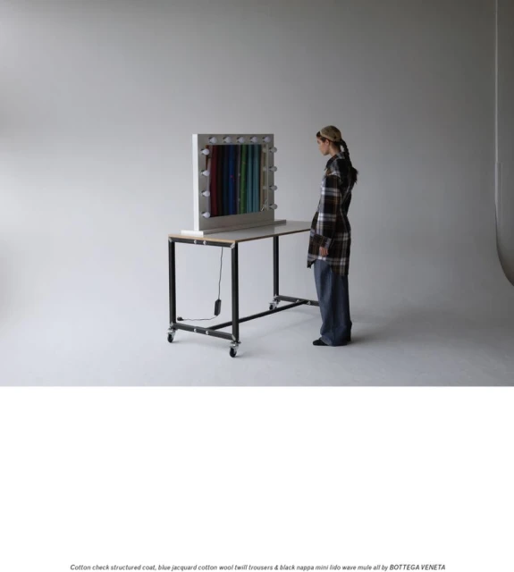 Woman in plaid coat and jeans stands near a table with colorful fabric display in a bright studio setting. Fashion editorial image.