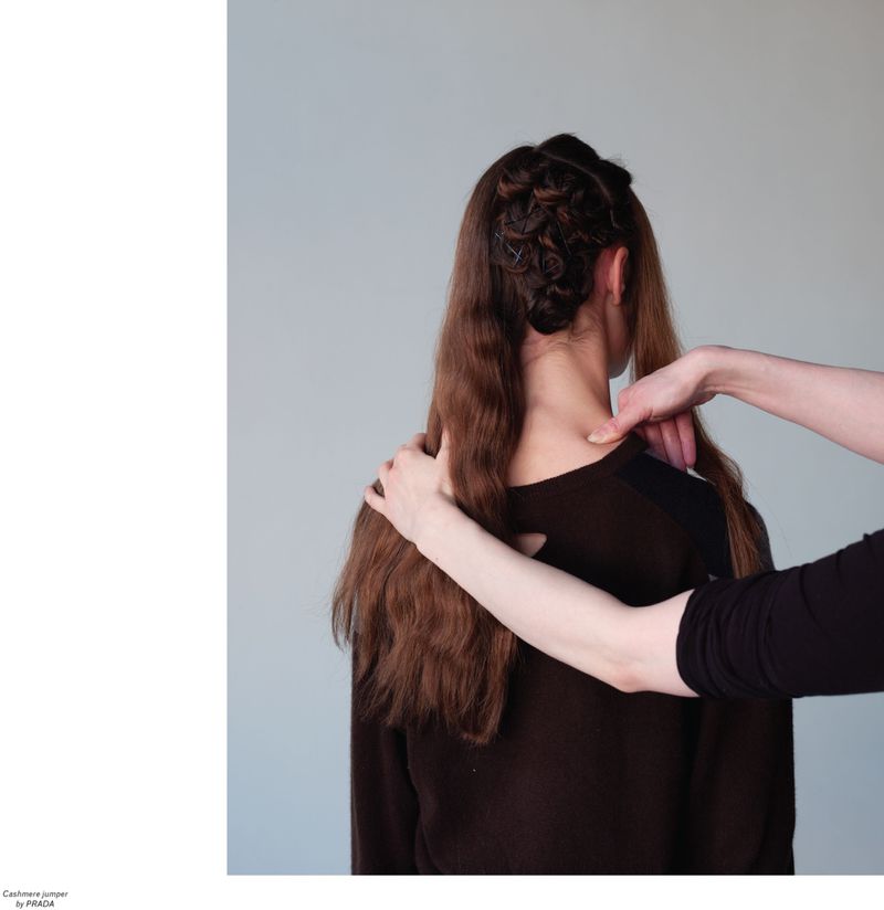 Back view of a woman with brown hair being styled, wearing a dark brown long-sleeved top against a neutral grey background, indoor setting.