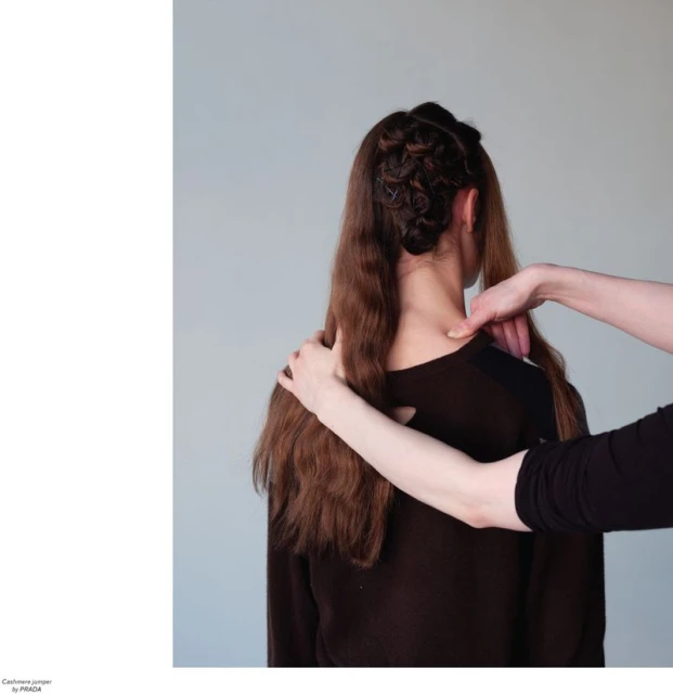Back view of a woman with brown hair being styled, wearing a dark brown long-sleeved top against a neutral grey background, indoor setting.