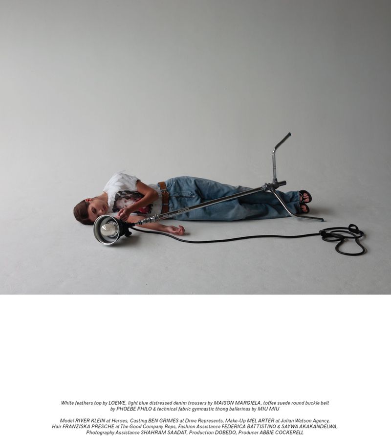 Young adult in light blue distressed denim lies next to a large film light in a studio with a simple white feather top and tan belt.