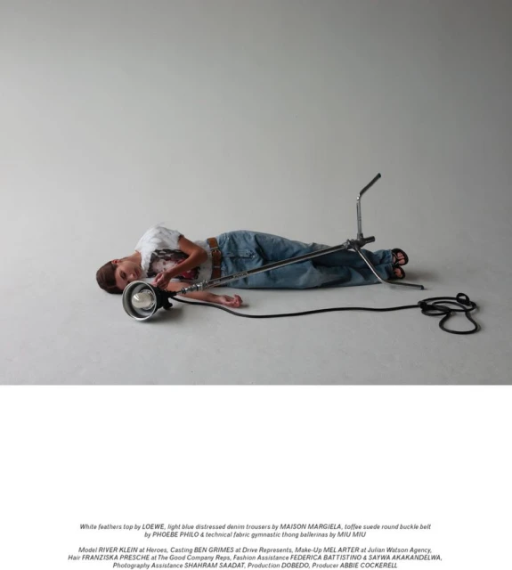 Young adult in light blue distressed denim lies next to a large film light in a studio with a simple white feather top and tan belt.
