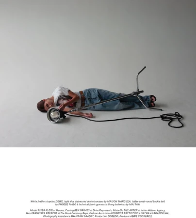 Young adult in light blue distressed denim lies next to a large film light in a studio with a simple white feather top and tan belt.
