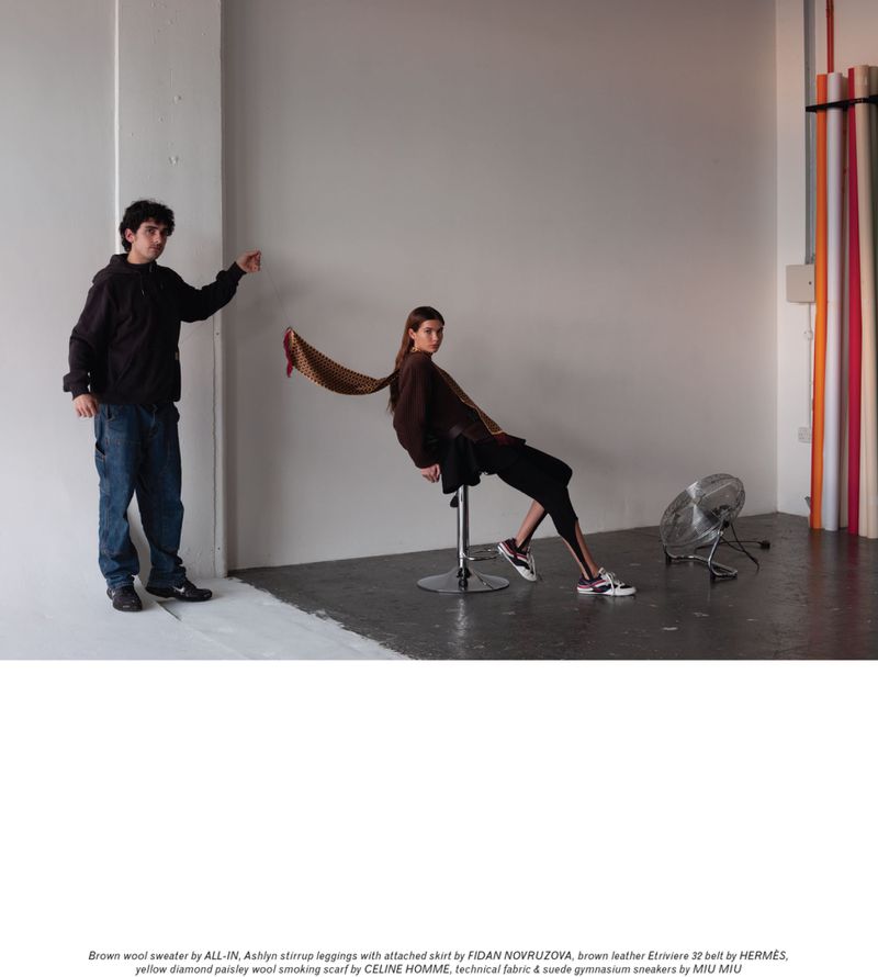 Fashion editorial shot of a woman seated on a stool with a scarf blowing behind her, and a man standing to her left in a studio setting.