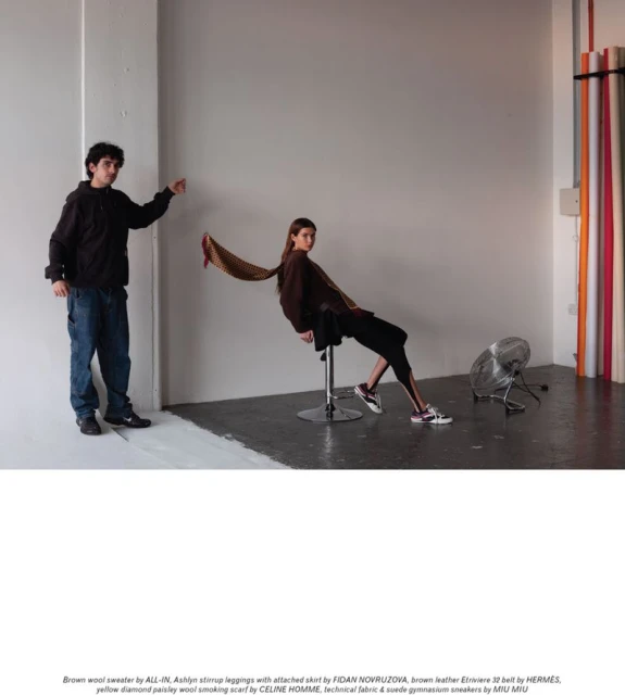 Fashion editorial shot of a woman seated on a stool with a scarf blowing behind her, and a man standing to her left in a studio setting.