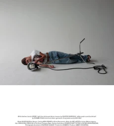Model River Klein lying on a grey seamless studio floor beside a metal light fixture with a coiled black cord.