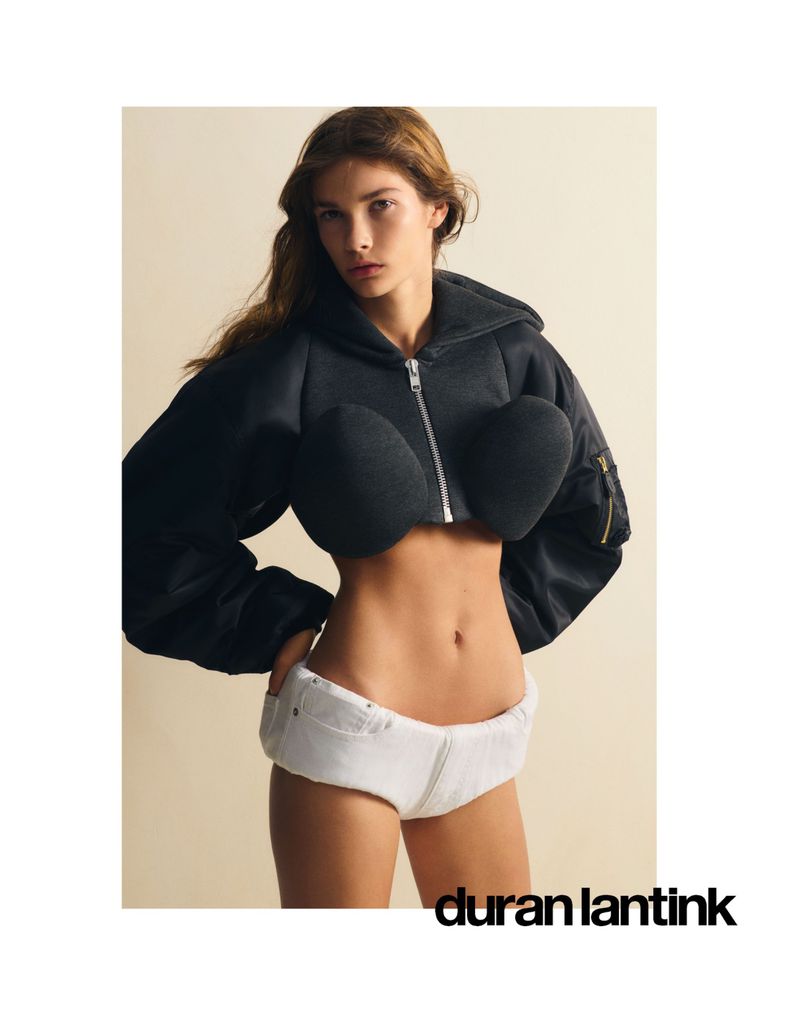 Model wearing a black zip-up cropped jacket with rounded structured chest pads and white shorts against a pale background