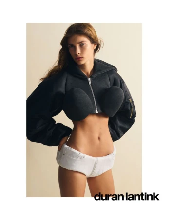 Model wearing a black zip-up cropped jacket with rounded structured chest pads and white shorts against a pale background