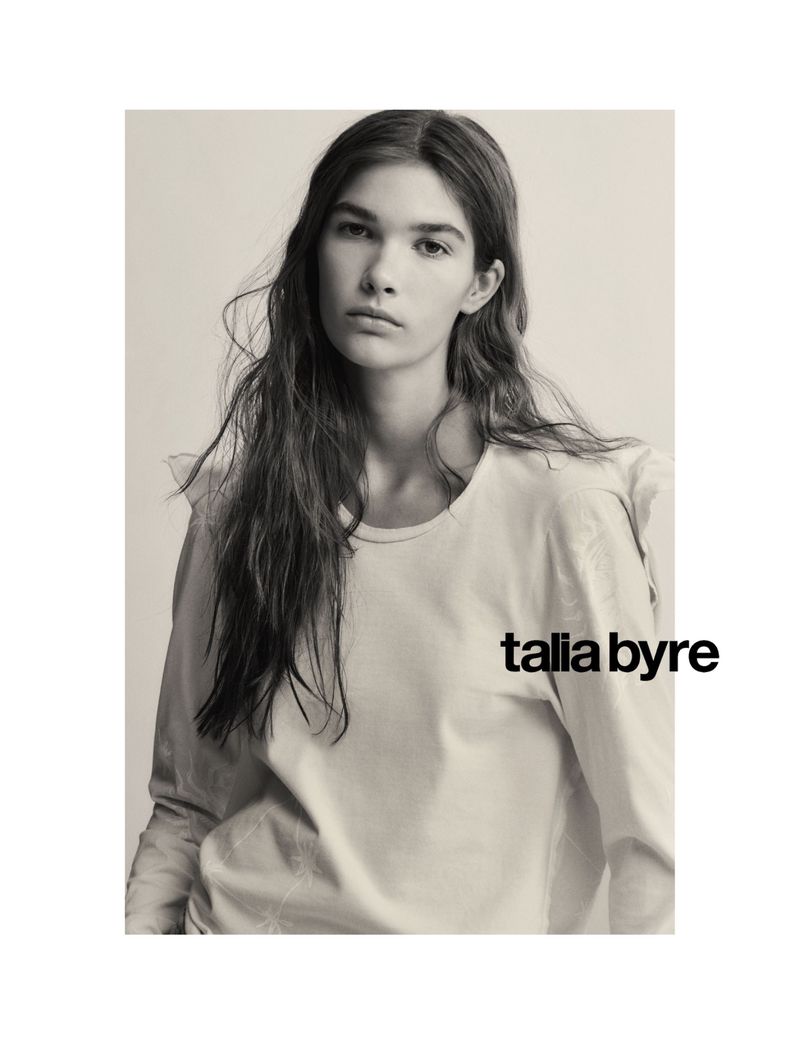 Monochrome portrait of a young woman with long hair wearing a light-colored long-sleeve shirt, posing in front of a neutral backdrop.