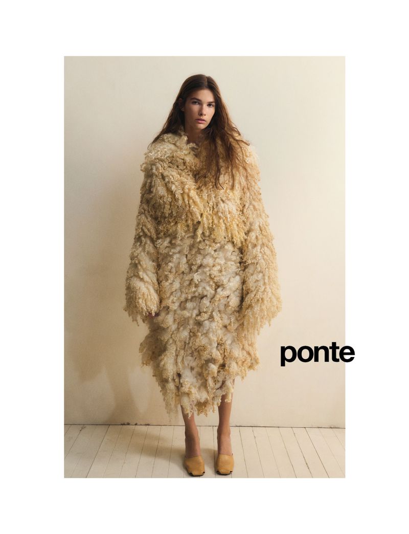 A young woman stands in a studio wearing an oversized, beige, textured, shaggy coat and low heel shoes against a neutral backdrop.