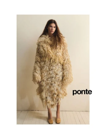 A young woman stands in a studio wearing an oversized, beige, textured, shaggy coat and low heel shoes against a neutral backdrop.
