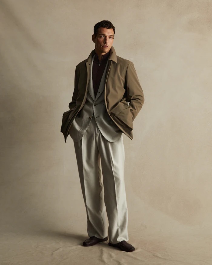 Full-length studio shot of an adult man in a grey suit and brown jacket, posing against a neutral backdrop with soft, diffused lighting.