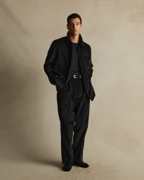 Man wearing a black jacket and trousers standing against a textured neutral background by photographer Rob Tennent
