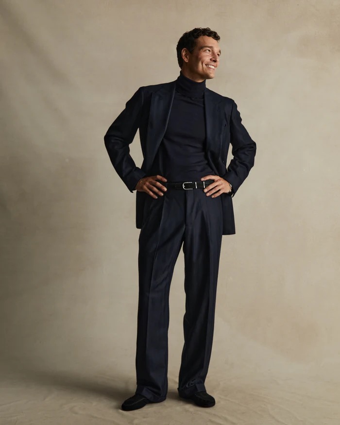 Full-length portrait of a smiling adult male with dark hair, wearing a dark suit with a turtleneck, standing indoors with hands on hips.