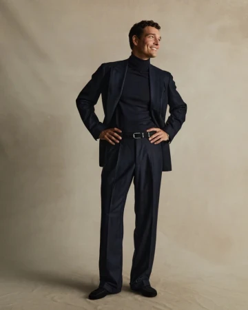 Full-length portrait of a smiling adult male with dark hair, wearing a dark suit with a turtleneck, standing indoors with hands on hips.