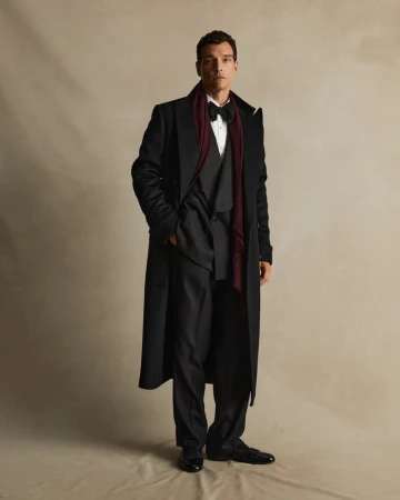 Man in black coat, dark suit, white shirt, black bowtie, and dark red scarf standing against a neutral studio backdrop.