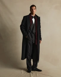 Man in black coat, dark suit, white shirt, black bowtie, and dark red scarf standing against a neutral studio backdrop.