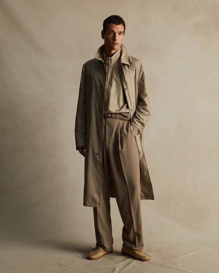 A man in a tan trench coat and matching pants and shoes poses in a studio setting with a neutral backdrop for a fashion shoot.