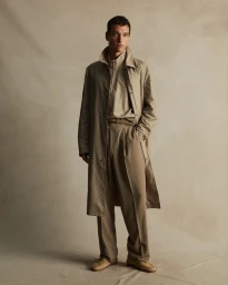Man wearing a beige trench coat and matching trousers standing against a neutral studio backdrop