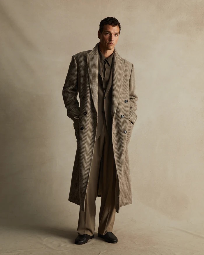 A stylish adult man in a herringbone overcoat, dress shirt, wide-leg pants, and loafers, captured in a neutral-toned photo studio.