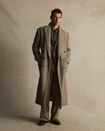 A stylish adult man in a herringbone overcoat, dress shirt, wide-leg pants, and loafers, captured in a neutral-toned photo studio.