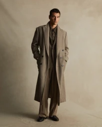 Man wearing a long patterned double-breasted coat and trousers standing against a textured neutral backdrop.