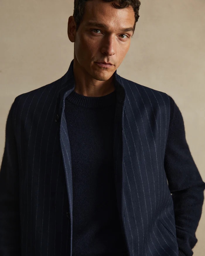 Portrait of a brunette adult male wearing a navy sweater and pinstripe vest looking at the camera in a neutral indoor setting.