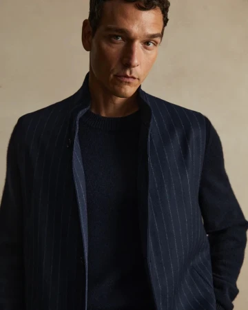 Portrait of a brunette adult male wearing a navy sweater and pinstripe vest looking at the camera in a neutral indoor setting.