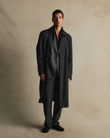Man wearing a dark trench coat over a patterned grey suit and black trousers against a neutral textured studio background.