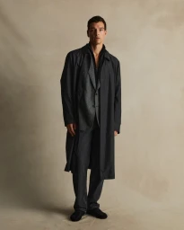 Man wearing a dark trench coat over a patterned grey suit and black trousers against a neutral textured studio background.