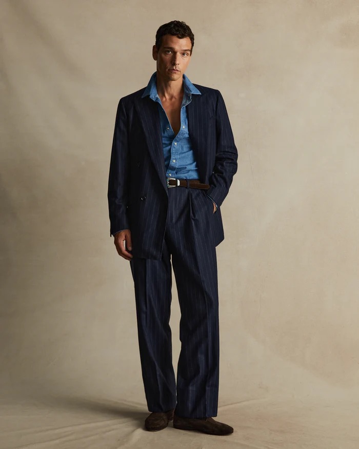 A man in a pinstripe suit stands against a neutral background, wearing a denim shirt and brown belt, showcasing a vintage-inspired fashion style.
