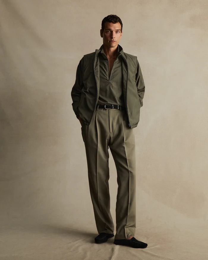 Full-length shot of a male model in a studio, wearing a green shirt, vest, and khaki pants against a neutral backdrop in a portrait.