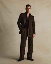 Man standing wearing a brown suit jacket, matching trousers, and a black zippered top against a neutral textured backdrop.