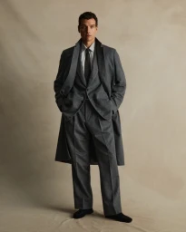 Man wearing a charcoal grey suit and matching overcoat standing against a neutral studio background.