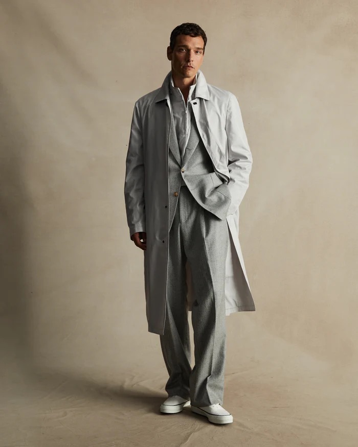 Full-length shot of a man in a grey three-piece suit and trench coat, posing in front of a neutral beige background, showcasing fashion.