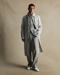 Man wearing a light grey trench coat and matching suit pants standing against a textured neutral backdrop.