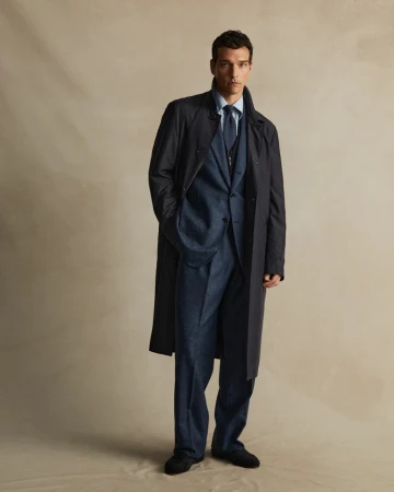 Man wearing a dark overcoat and blue suit standing against a textured backdrop