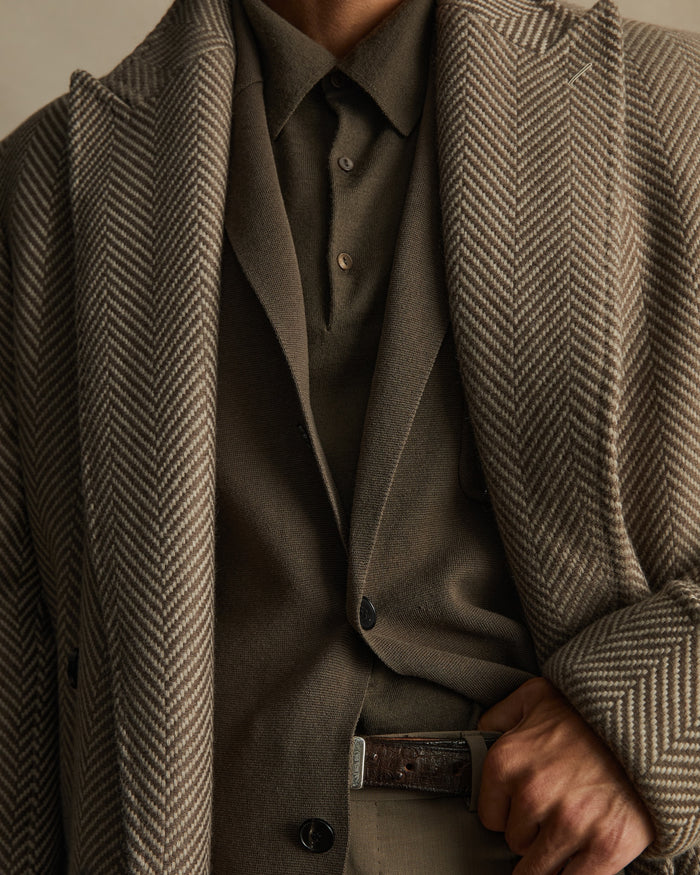 Adult male wearing a herringbone coat and a brown suit, posing indoors with a buttoned collared shirt and a leather belt.