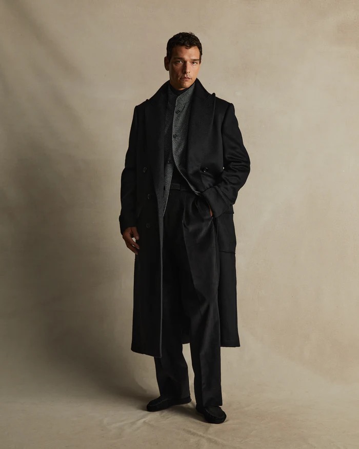 A single adult male poses in an indoor setting, wearing a full-length black coat, dress pants and black shoes. Neutral background. 