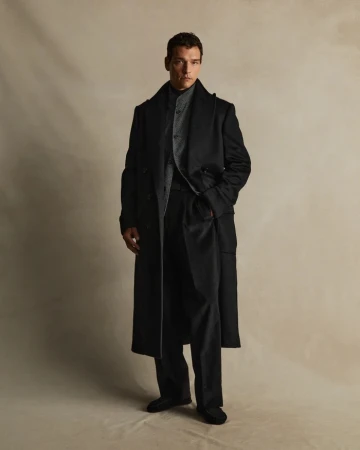 A single adult male poses in an indoor setting, wearing a full-length black coat, dress pants and black shoes. Neutral background. 