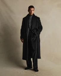 Man wearing a long black coat over a grey patterned vest against a textured neutral background