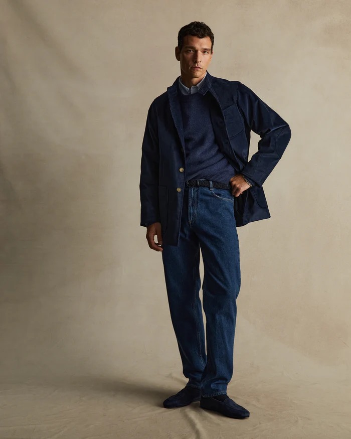A full shot of a man in a navy corduroy jacket and jeans poses against a neutral backdrop in a studio fashion shot.