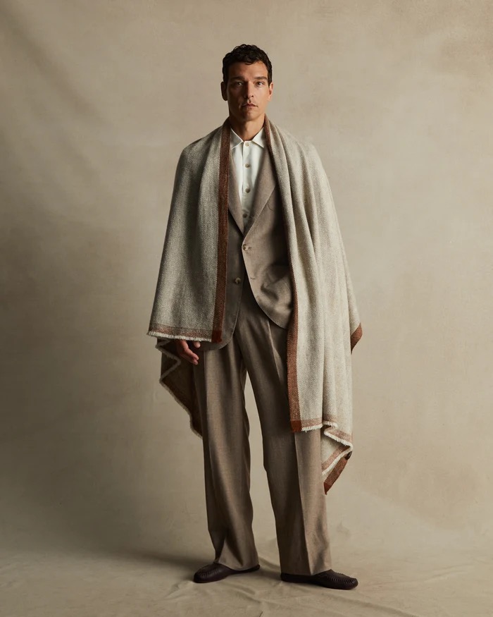 Man in a tan suit and black shoes standing indoors with a beige shawl. Neutral color palette, full shot. Fashion editorial.