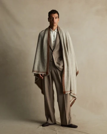 Man in a tan suit and black shoes standing indoors with a beige shawl. Neutral color palette, full shot. Fashion editorial.