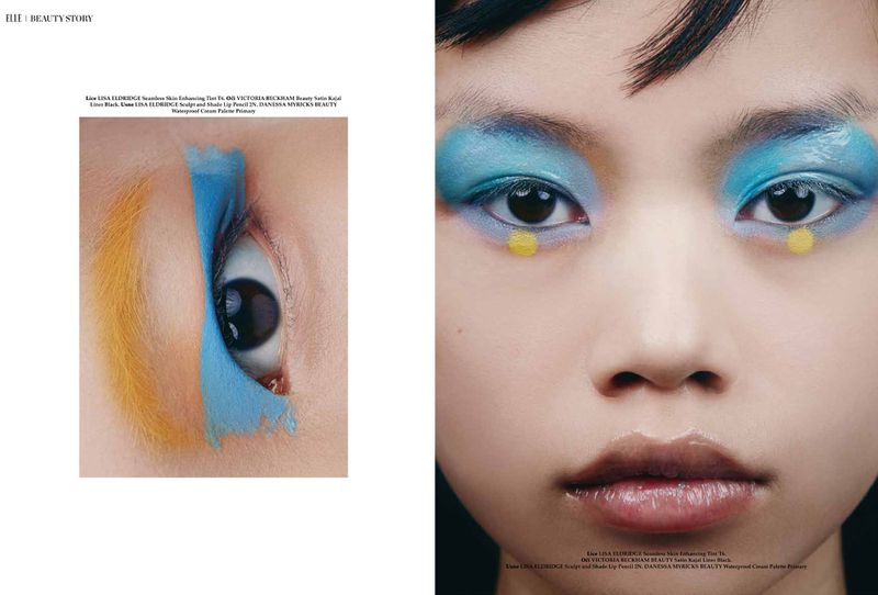 Close-up fashion shot of an Asian woman with blue eyeshadow, yellow face dots, glossy lips, and dramatic eye makeup. Beauty editorial.