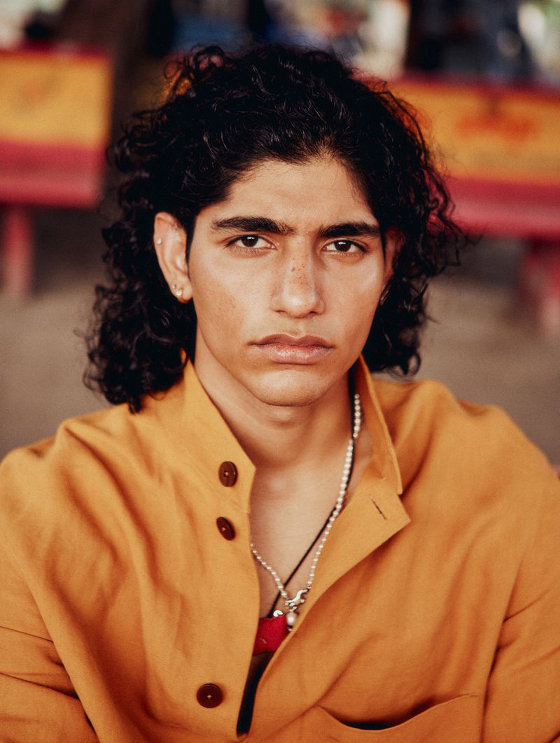 Portrait of a young adult man with dark curly hair, wearing an orange button-down shirt and a pearl necklace with pendants.