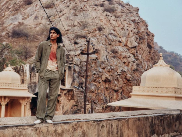 Young man poses outdoors wearing a sage green linen suit in front of a stone wall, domed structure, and rocky mountain backdrop.