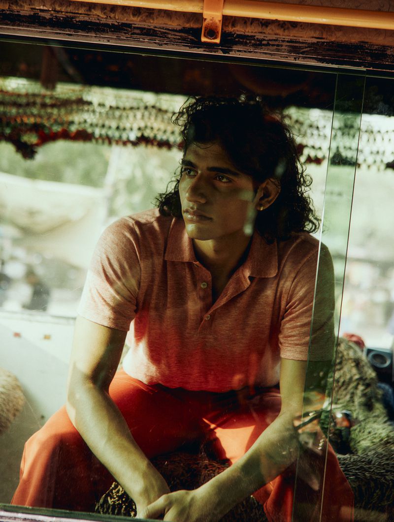 Portrait of a dark-haired young adult male in a salmon-colored polo and coral pants, sitting inside what appears to be an auto rickshaw.