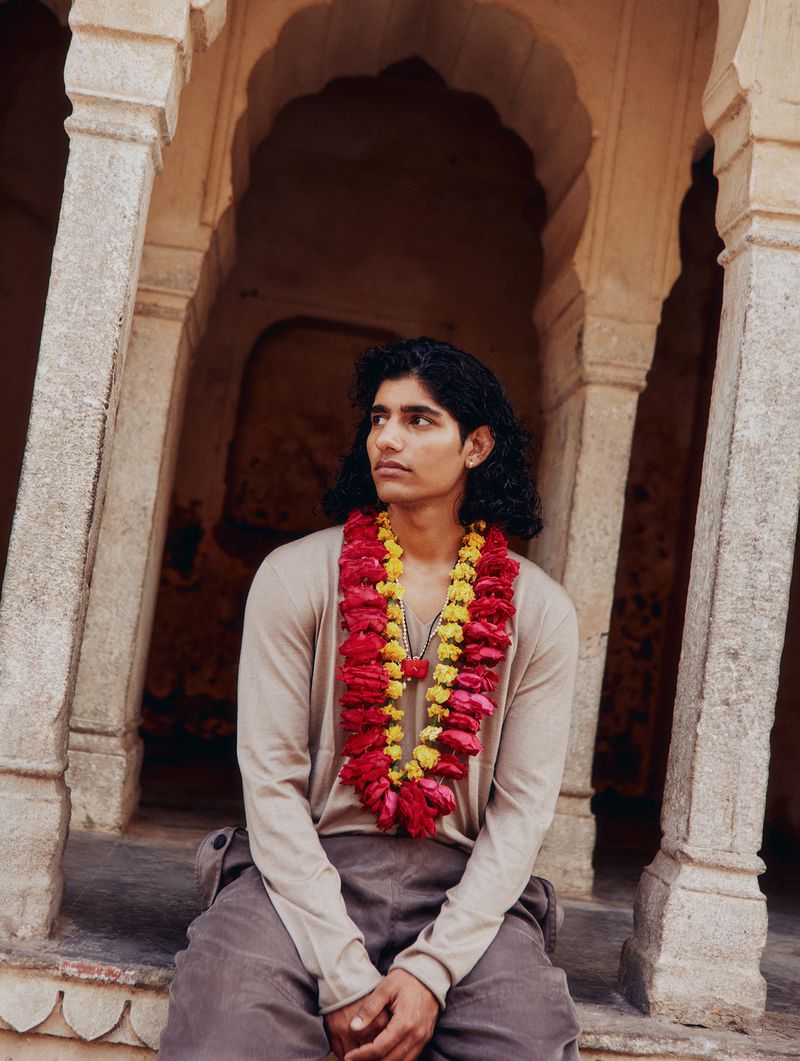 Young adult sits in a tan stone structure wearing a flower garland and looking to the left, set against a neutral background.