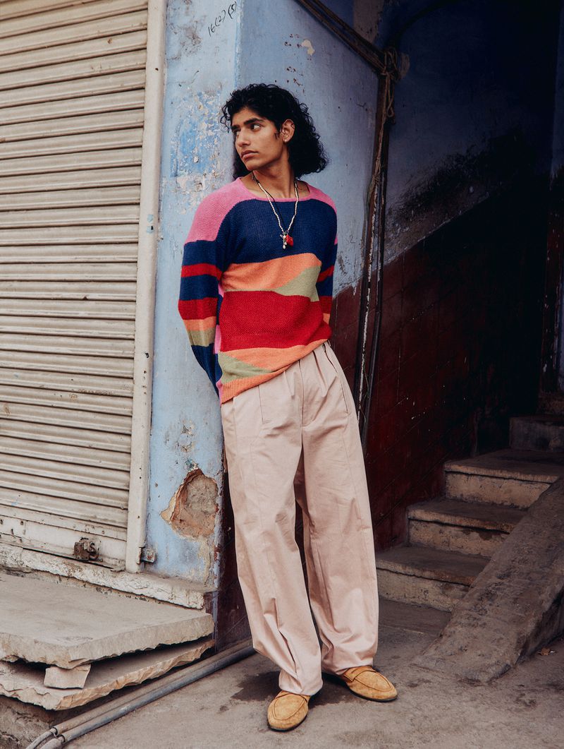 A young adult male in a colorful striped sweater and loose-fitting pants leans against a weathered wall, posing on an urban street.