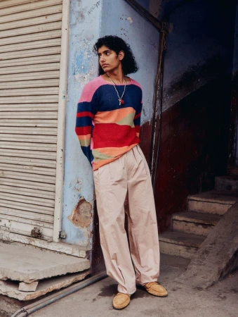 A young adult male in a colorful striped sweater and loose-fitting pants leans against a weathered wall, posing on an urban street.